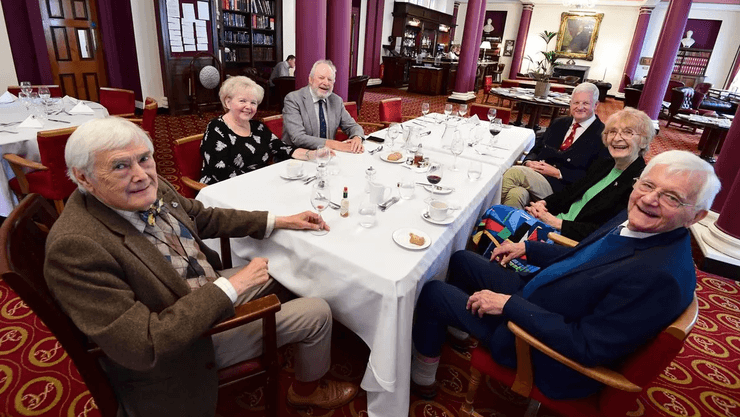 Группа пожилых людей сидит за большим накрытым столом в ресторане Атенеума, улыбаясь и общаясь