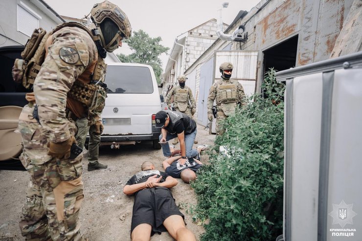 Вооружённые правоохранители в тактическом снаряжении и сотрудник полиции проводят задержание нескольких мужчин возле промышленного здания