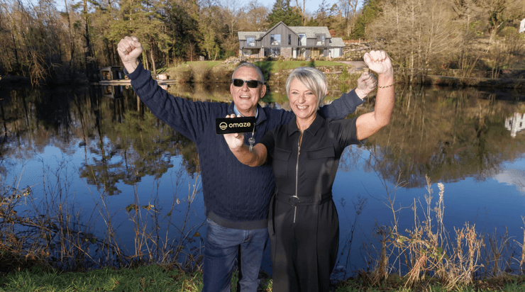 Супружеская пара с ключом от выигранного дома стоит на берегу частного озера