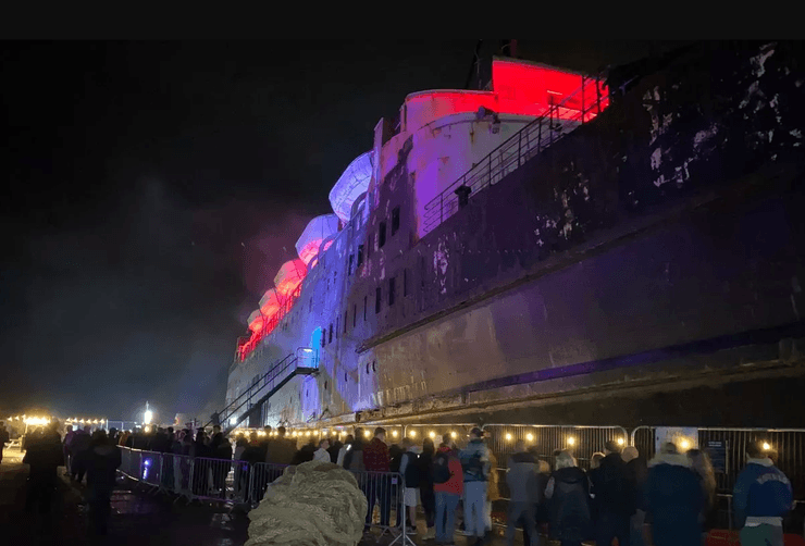 Толпа людей у ржавого борта корабля "Герцог Ланкастер", подсвеченного красным и синим светом