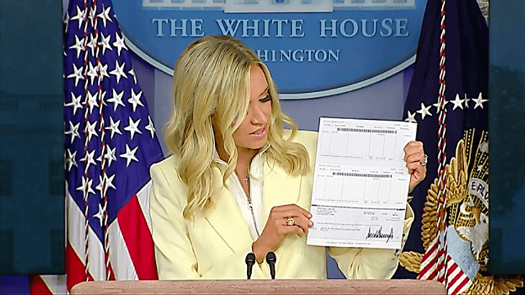 Пресс-секретарь Белого дома Кейли Макинани показывает чек на $100 тыс., пожертвованные Трампом Минздраву США