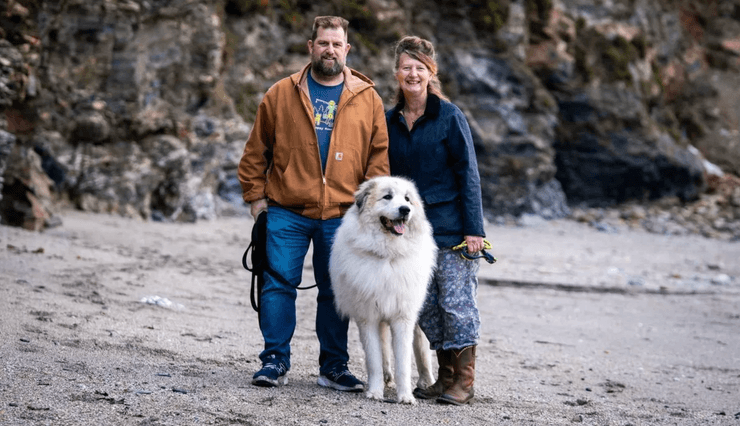 Величезний білий піренейський гірський пес стоїть поруч із чоловіком і жінкою на кам'янистому пляжі, вражаючи своїми розмірами