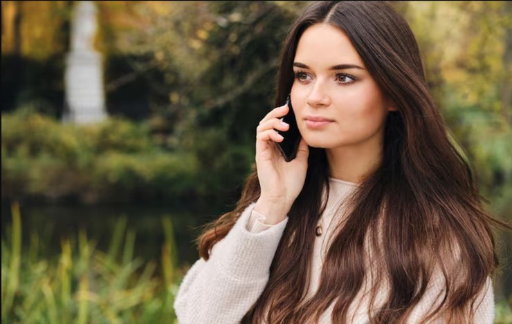 Молодая женщина с длинными тёмными волосами говорит по телефону, стоя в парке