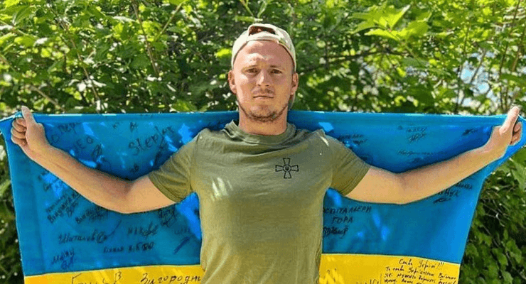 Український футболіст Роман Зозуля позує з прапором України, підписаним військовослужбовцями