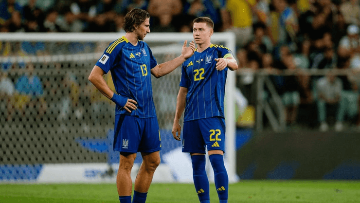 Два футболиста сборной Украины в синих формах обсуждают игру на поле во время матча, на заднем плане видны болельщики