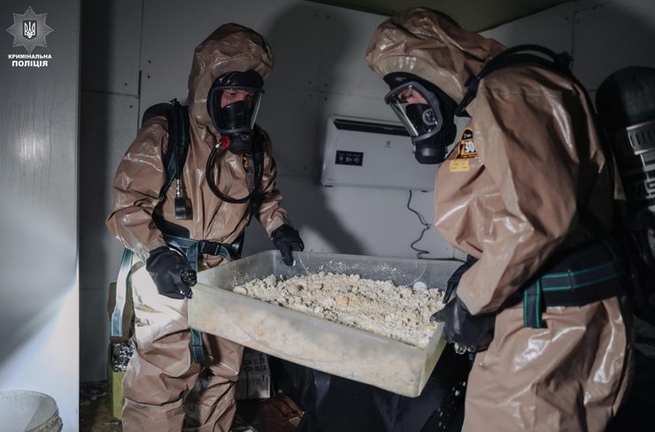 Двое сотрудников криминальной полиции в защитных костюмах и противогазах держат большой контейнер с белым комковатым веществом