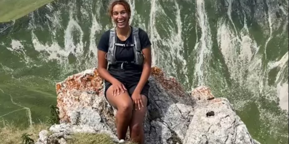 Молодая женщина улыбается, сидя на скале, рядом с ней ходят дружелюбные козы