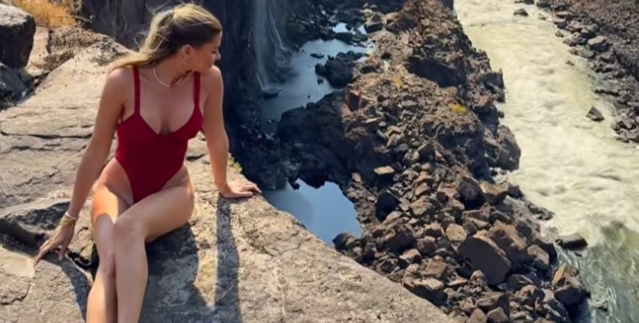 Жінка в червоному купальнику сидить на краю скелі біля водоспаду Вікторія, не підозрюючи, що за її спиною повзає змія