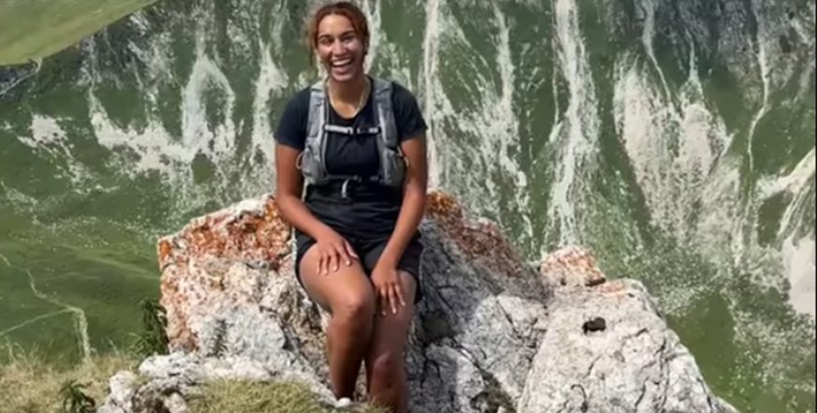 Молода жінка посміхається, сидячи на скелі, поруч із нею ходять доброзичливі кози