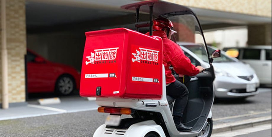 Курьер в красной форме Demae-can доставляет еду на трехколесном скутере в Японии