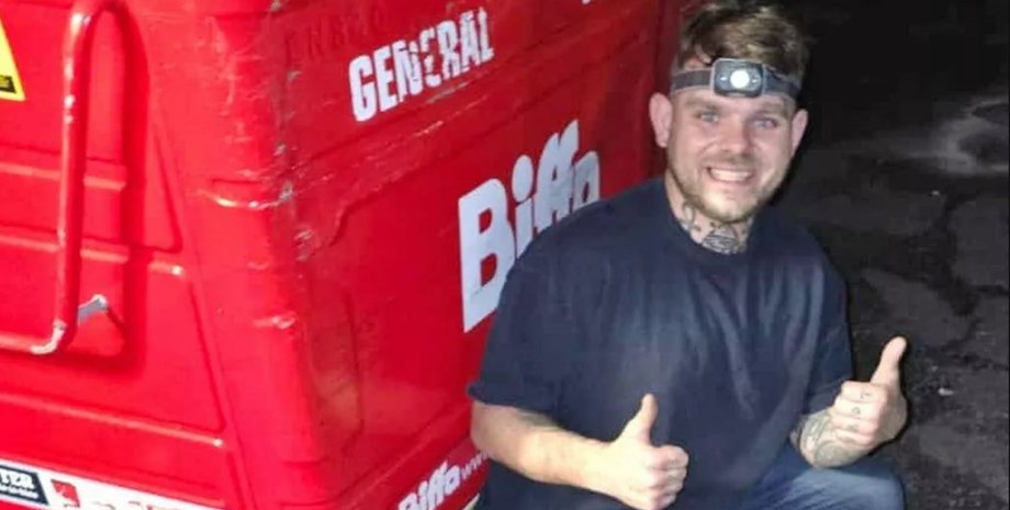 Чоловік, який займається пошуком викинутих речей, посміхається з великим червоним сміттєвим контейнером увечері