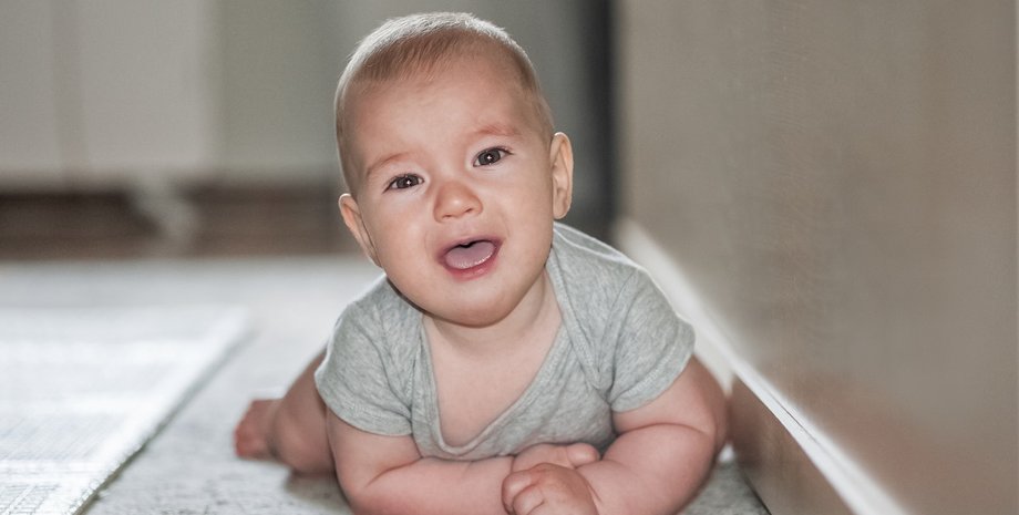 Младенец в серой футболке ползает по полу и смотрит в камеру