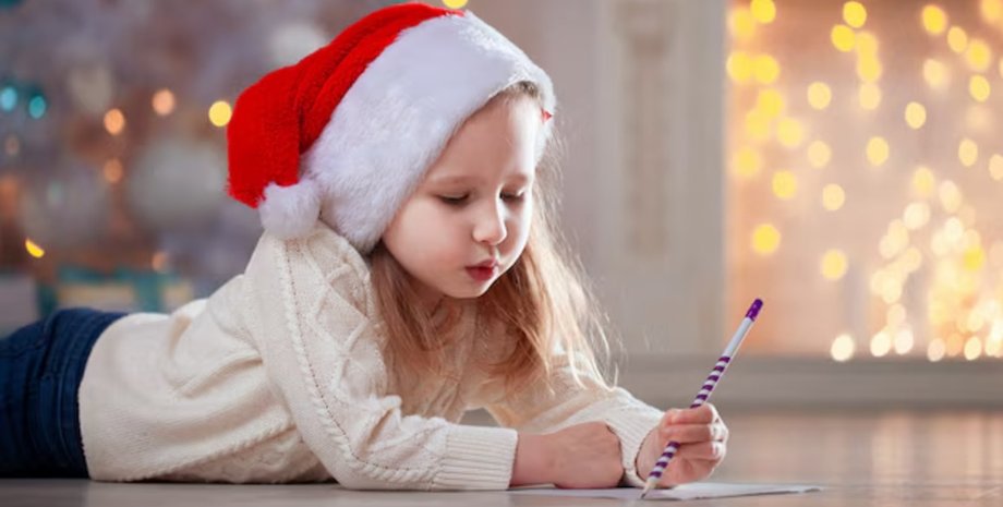 Маленькая девочка в красной шапке Санты лежит на полу и пишет рождественское письмо