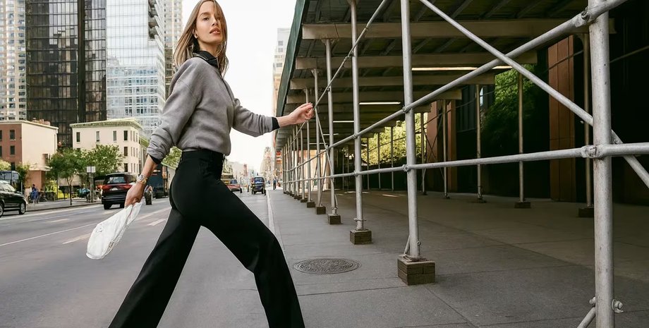 Молодая женщина в черных брюках и сером свитере идёт по улице города