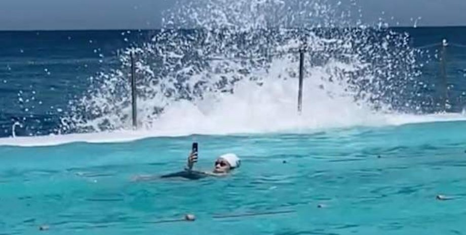 Женщина в шапочке плавает в бассейне Bondi Icebergs в Сиднее, держа телефон над водой