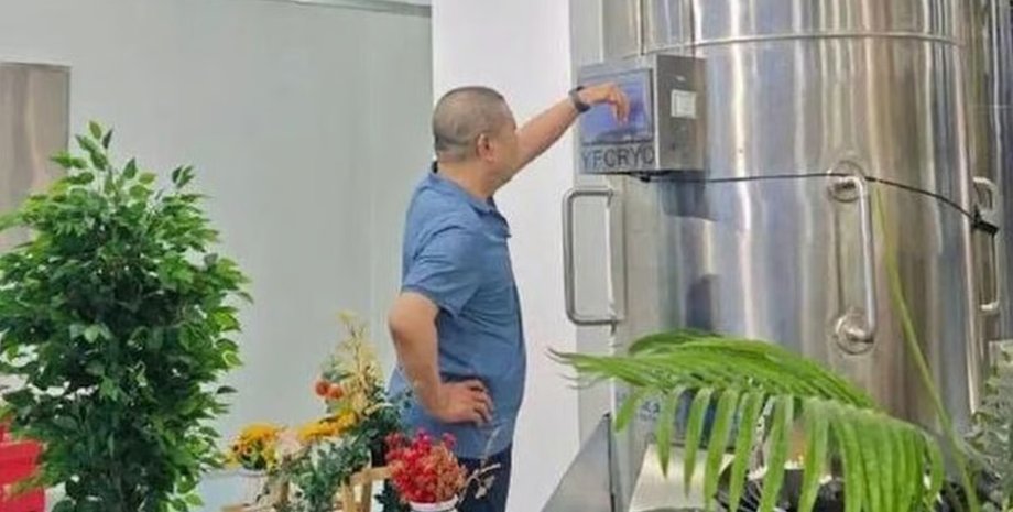 Чоловік у блакитній сорочці стоїть біля великої металевої кріокамери і натискає на панель управління