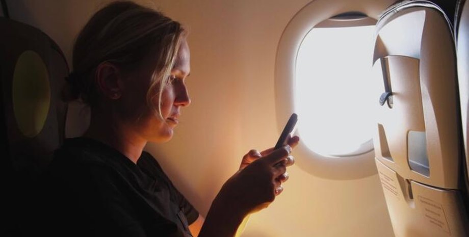 Жінка сидить біля ілюмінатора літака і дивиться в телефон