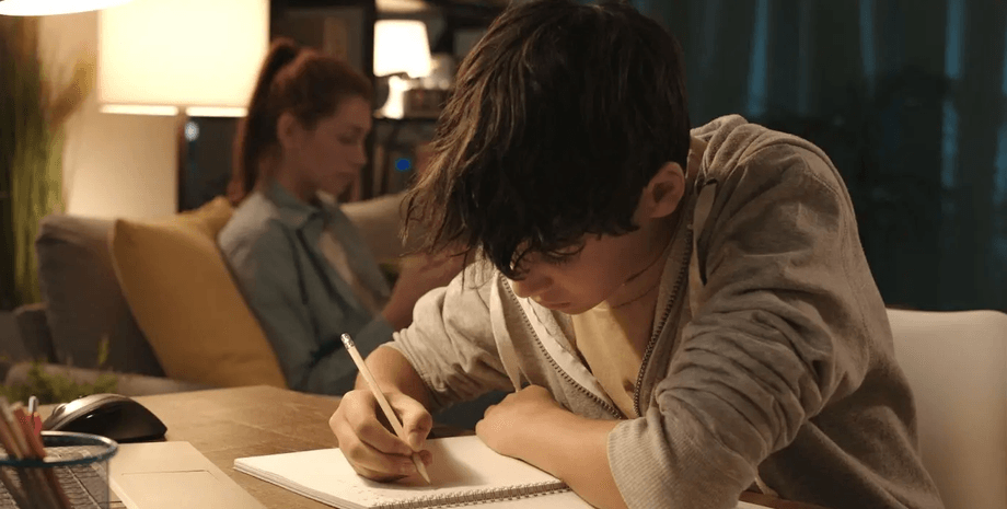 Мальчик усердно пишет в тетради за рабочим столом, рядом сидит женщина с телефоном