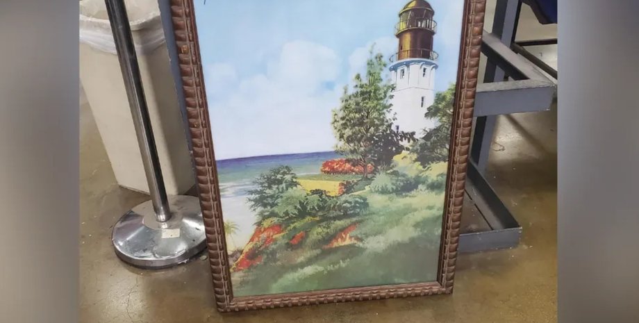Картина із зображенням маяка і морського узбережжя, що стоїть на підлозі в секонд-хенді, без видимої ціни на полотні.