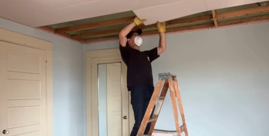 Чоловік на сходах знімає панелі підвісної стелі в спальні, виявляючи прихований простір