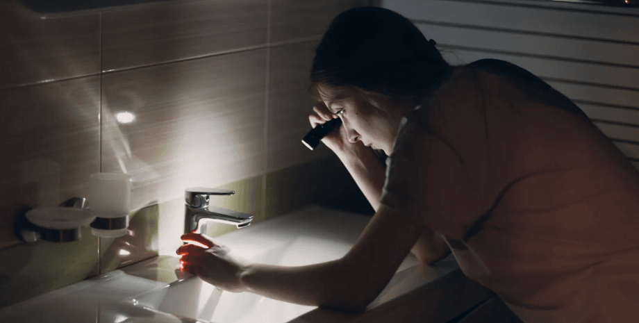 Женщина с фонариком наклоняется над раковиной в темной ванной комнате, проверяя источник странного шума посреди ночи