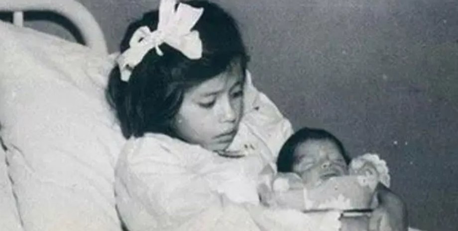 Дівчинка сидить на лікарняному ліжку і тримає на руках новонароджену дитину, загорнуту в ковдру