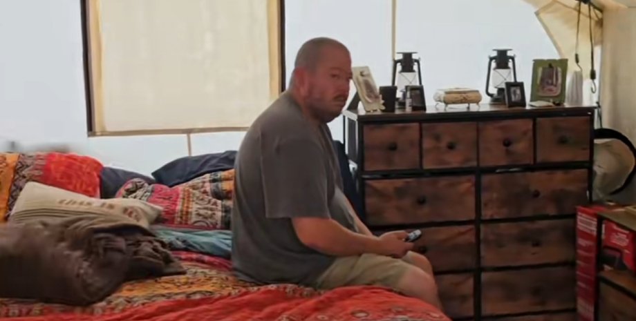 Чоловік сидить на ліжку всередині брезентового намету з дерев'яним комодом і домашнім інтер'єром