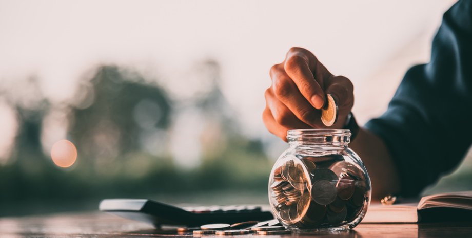 Рука людини опускає монету в скляну банку з накопиченнями