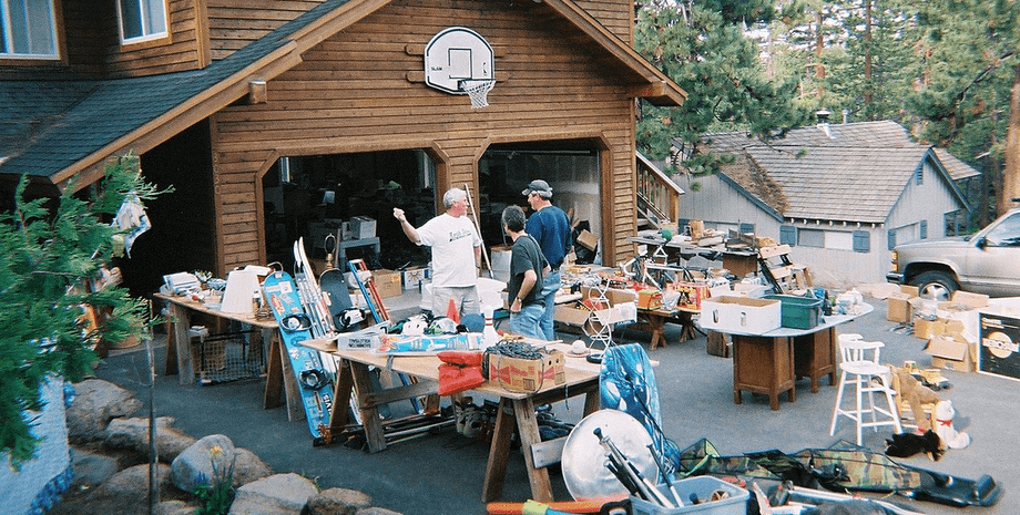 Люди осматривают вещи на гаражной распродаже среди столов с мебелью, коробками и предметами быта