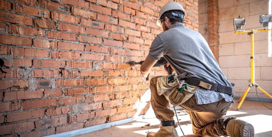 Мужчина в каске и рабочей одежде с инструментами демонтирует кирпичную стену в доме, готовя помещение к ремонту
