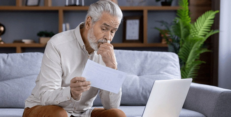 Літній чоловік сидить удома з документами і ноутбуком, вивчаючи папери і виглядаючи стурбованим
