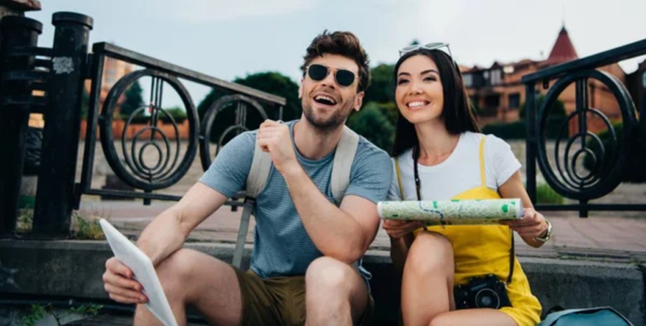 Щаслива пара туристів із картою під час подорожі Молода пара туристів сидить на вулиці з картою і планшетом, плануючи маршрут під час поїздки