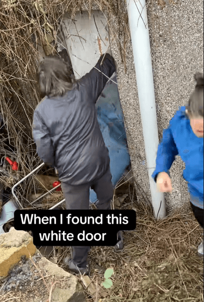 Дві жінки прибирають зарості та садове сміття, щоб відкрити старі білі двері, заховані за кущами