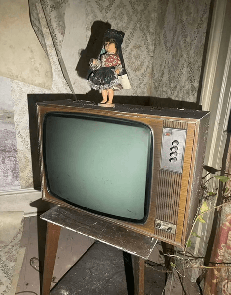 Старый коробочный телевизор 1970-х с куклой на фоне выцветших обоев в заброшенном доме в Уигане